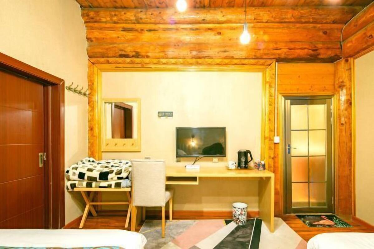 A room with a wooden ceiling, a desk with a television, a chair, a bed, and a door.