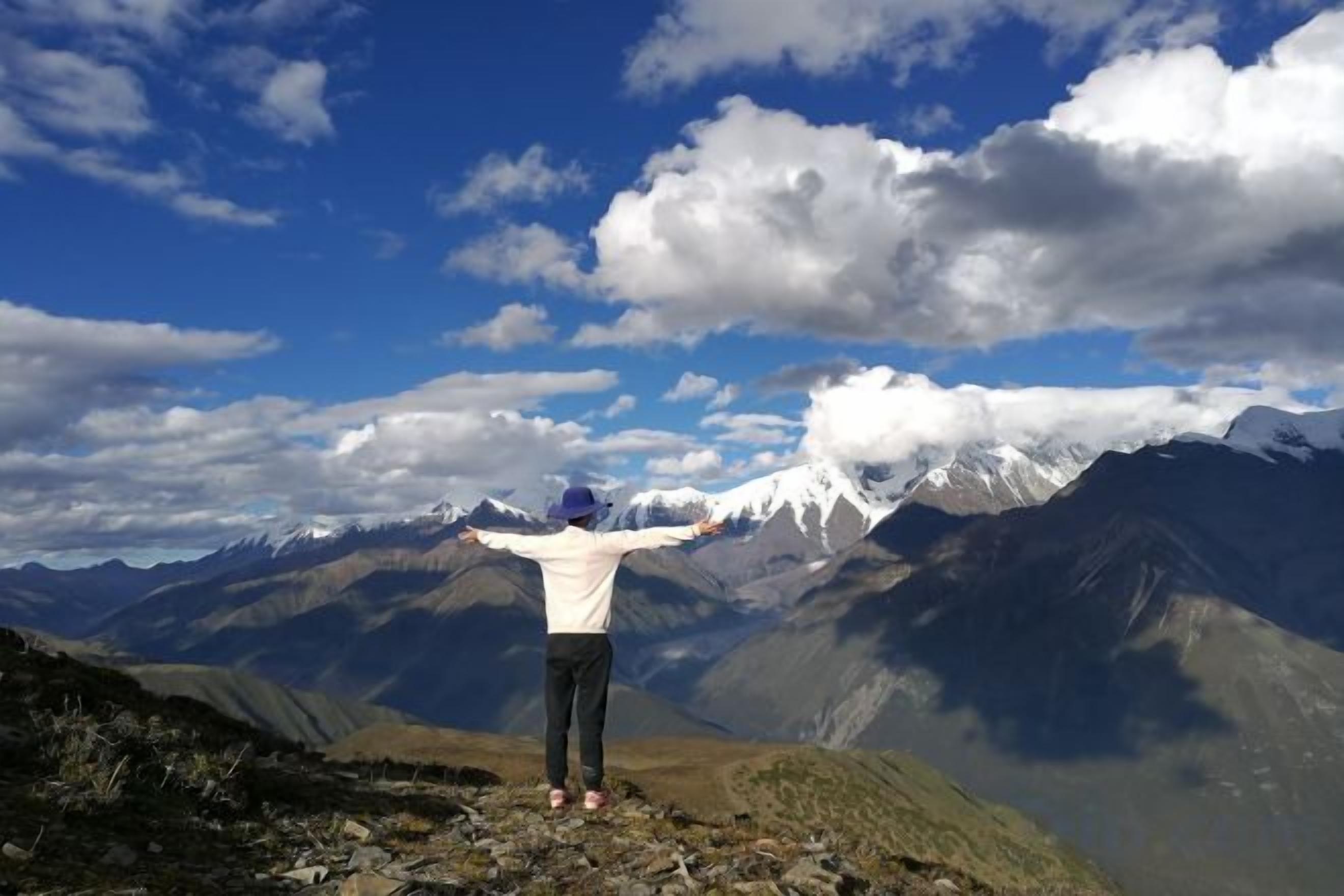 A person standing on a rocky outcrop with arms outstretched, overlooking a mountainous landscape with snow-capped peaks under a partly cloudy sky.