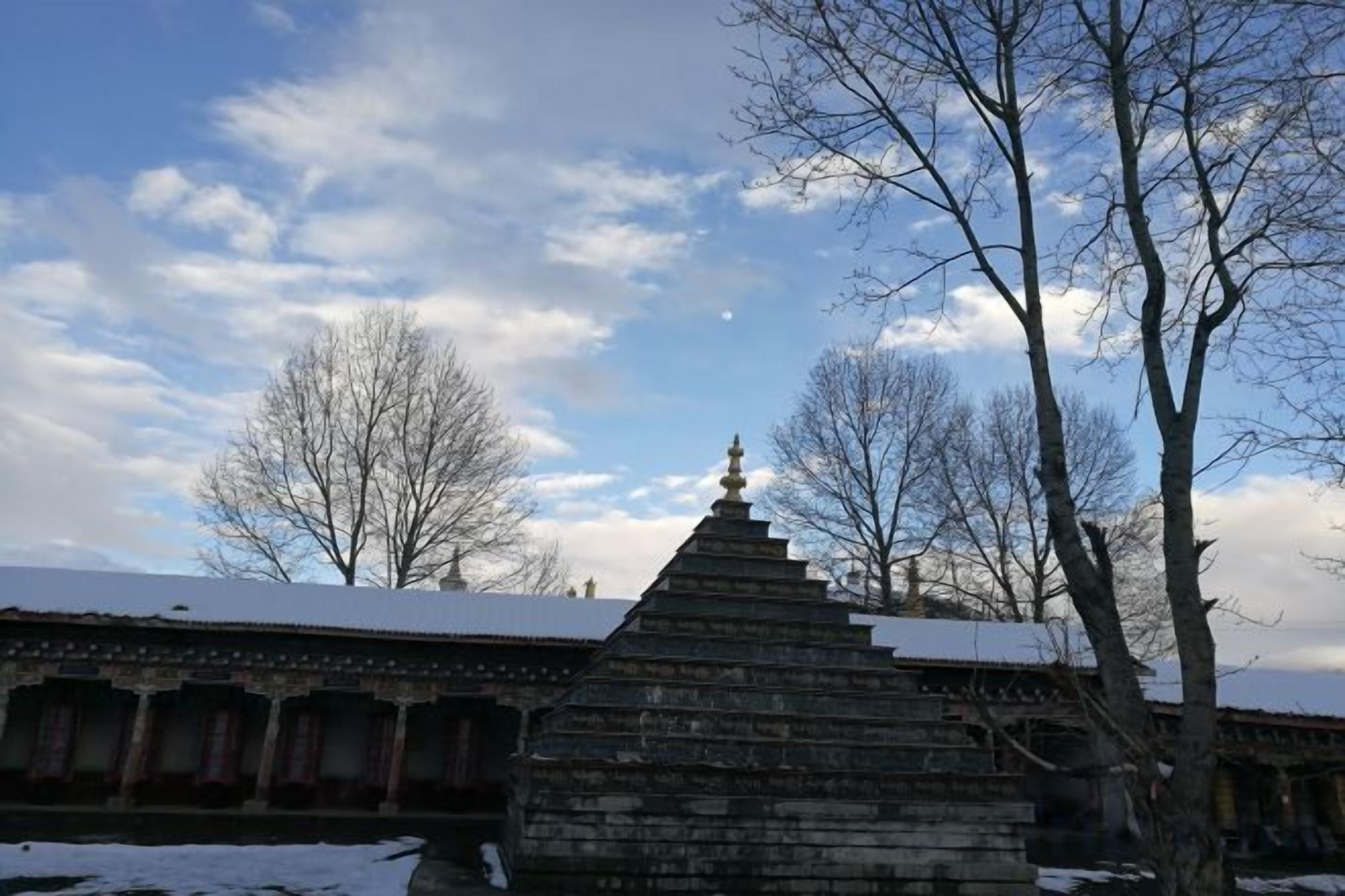 A stone structure with a tiered roof, possibly a monument or shrine, in front of a building with a snow-covered roof.