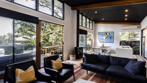 Un vaste espace de vie comprenant un canapé noir, deux fauteuils et une table à manger en bois. De grandes fenêtres offrent une vue sur l’extérieur.