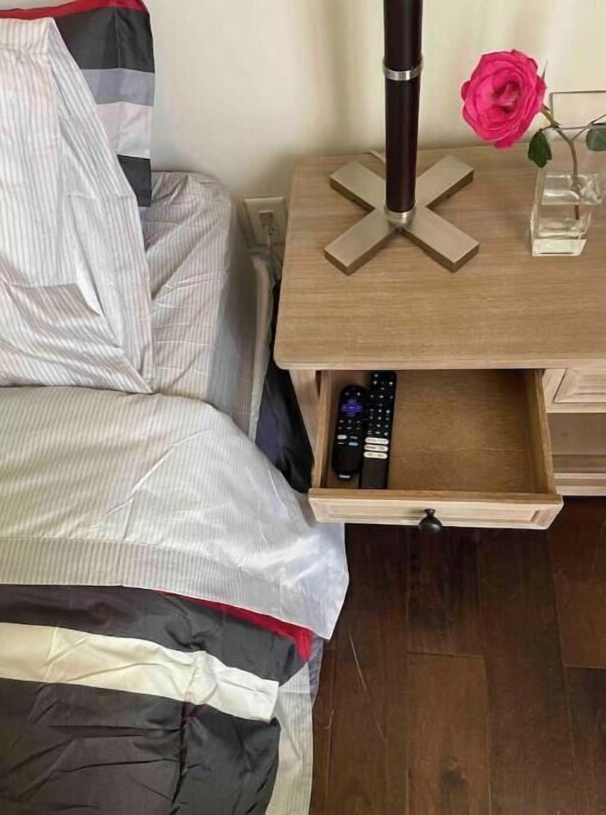 A bedroom with a bed, a bedside table with a remote control drawer, a vase with a single rose, and a black cylindrical object on the table.