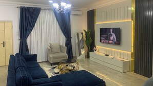 A modern living room with a sofa set, a coffee table, a TV mounted on the wall, and a decorative plant. - 4mins to Aso Rock Villa, King Bed, Chef, Free Cleaning, 24/7 Electricity, Games (Abuja)