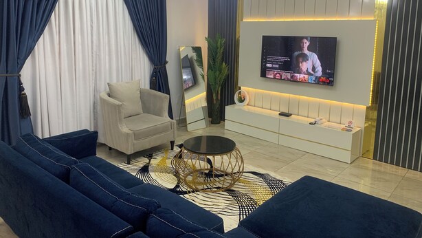 A modern living room with a sofa set, a coffee table, a TV mounted on the wall, and a chandelier. - 4mins to Aso Rock Villa, King Bed, Chef, Free Cleaning, 24/7 Electricity, Games (Abuja)
