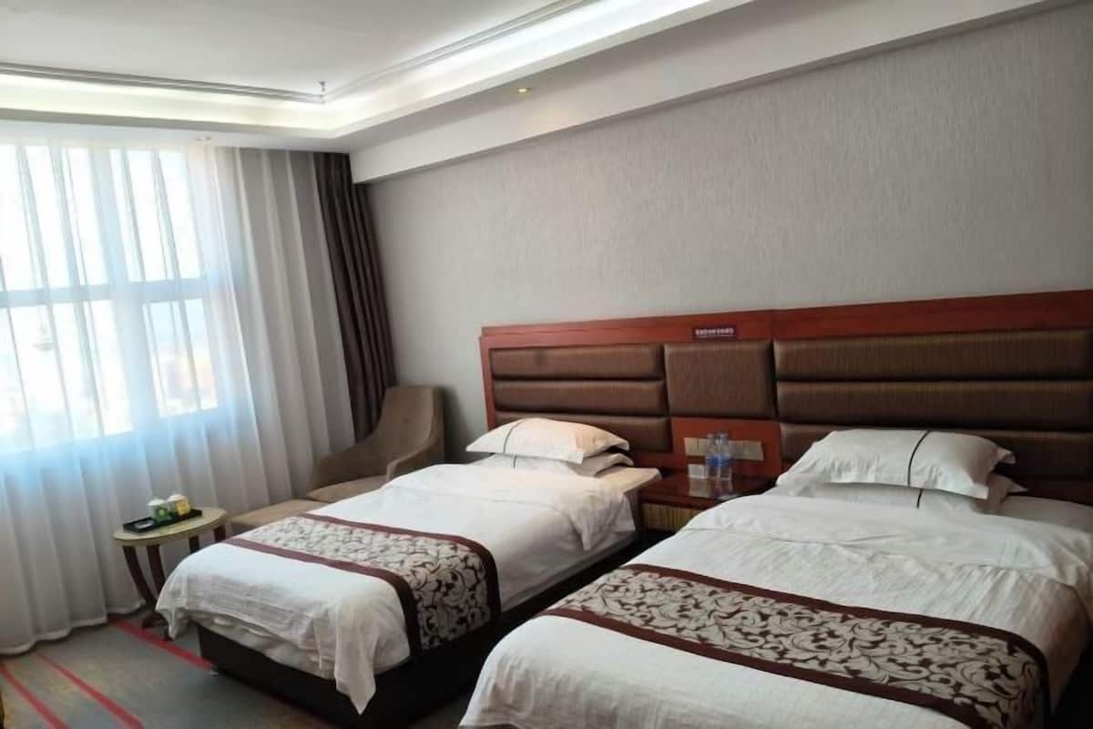 A hotel room with two beds, a chair, a small table with a phone and a bottle, and a window with curtains.