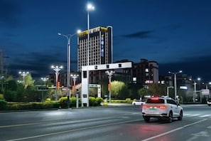 Een nachtelijk stadsbeeld met een witte auto, straatverlichting en gebouwen op de achtergrond.