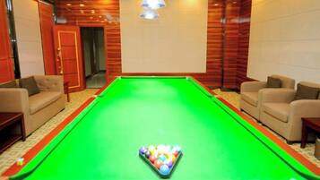 A pool table with a cue ball, surrounded by four armchairs, in a room with wooden paneling and a chandelier.