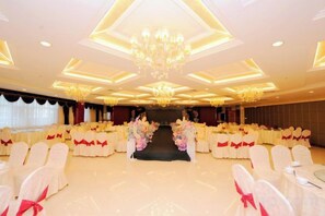 A banquet hall with rows of white chairs, a stage with floral arrangements, and a chandelier.
