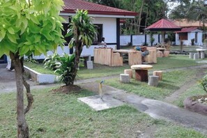 Un espacio al aire libre con mesas y sillas, rodeado de follaje y un pequeño edificio con techo rojo.