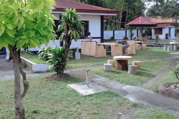 An outdoor seating area with tables and chairs, surrounded by greenery and a small building with a red roof.