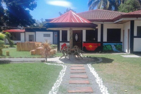 Un edificio con techo rojo, un camino pavimentado que lleva a la entrada y un pequeño jardín con muebles de madera.