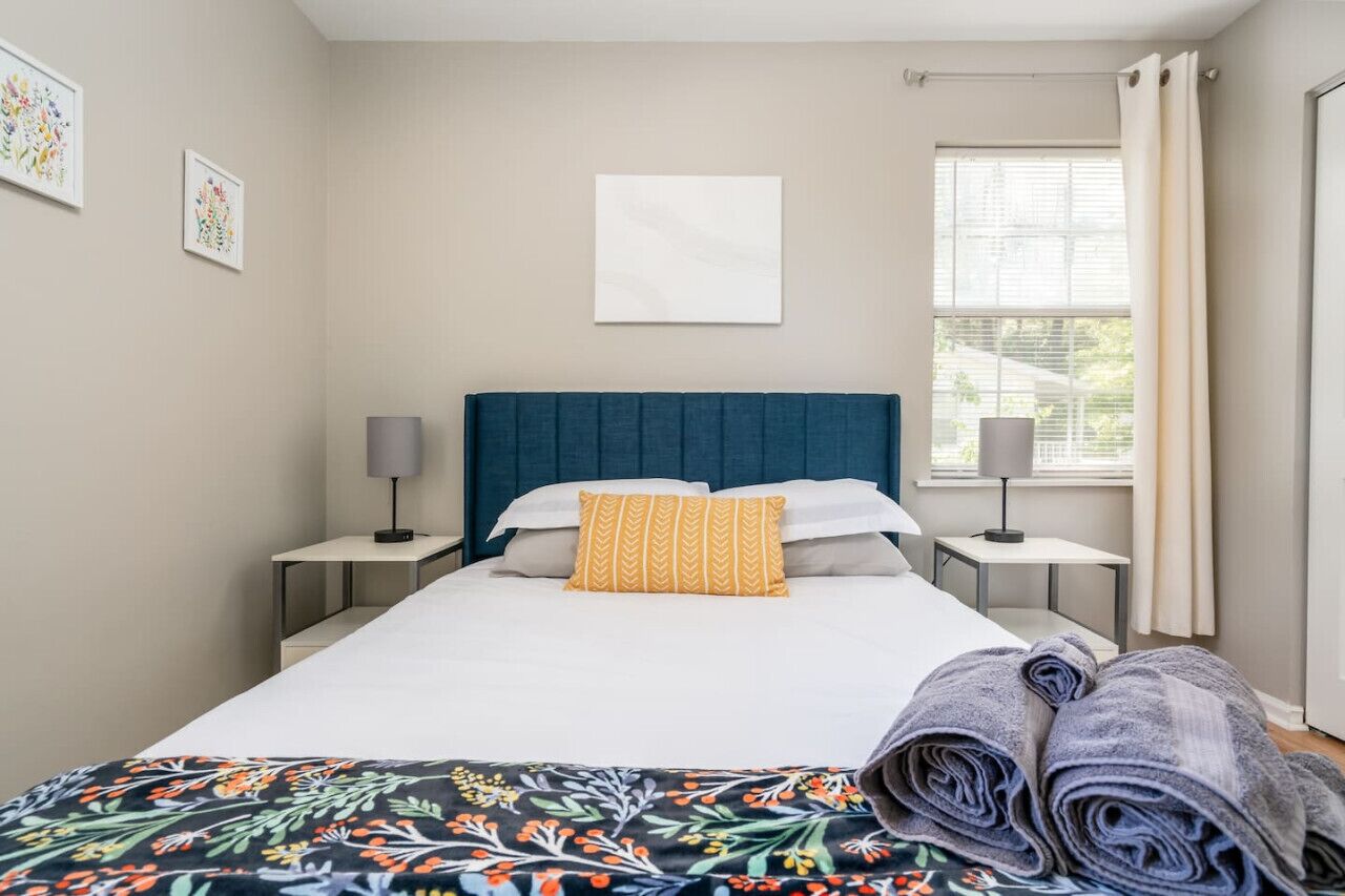 A neatly made bed with a blue headboard, white bedding, and a floral-patterned blanket. There are two bedside tables with lamps, a window with curtains, and a wall-mounted artwork.