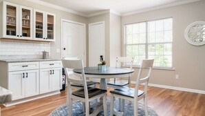Een keuken met witte kasten, een ronde tafel en stoelen. Er is een raam met zonwering en een decoratief wandobject.