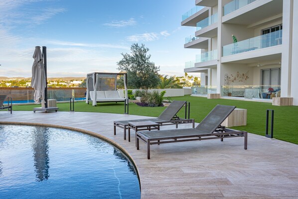 Un área junto a la piscina con sillones de descanso, un elemento decorativo de agua y un edificio moderno con balcones.