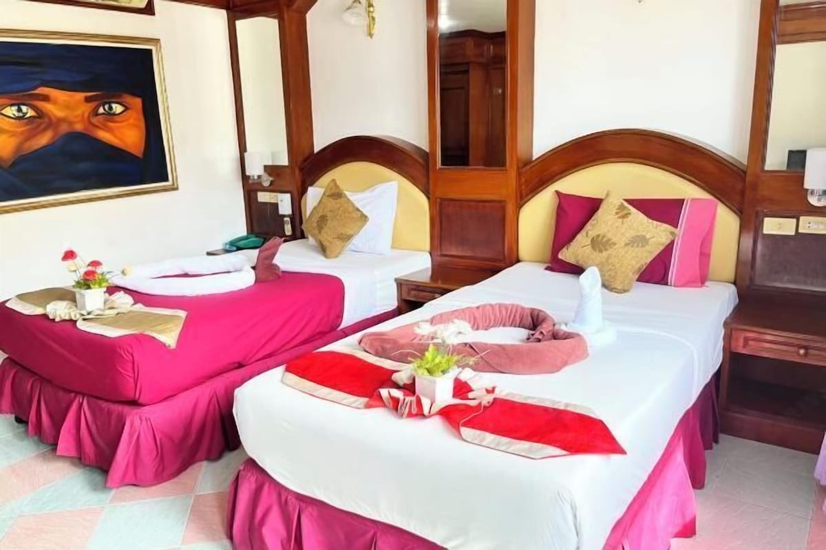 Two single beds with wooden headboards, a painting on the wall, and a small table with a vase of flowers.