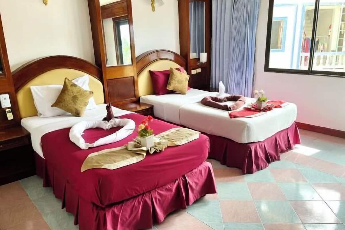 Two double beds with red bedding, a wooden headboard, a small table with a vase, and a window with curtains.