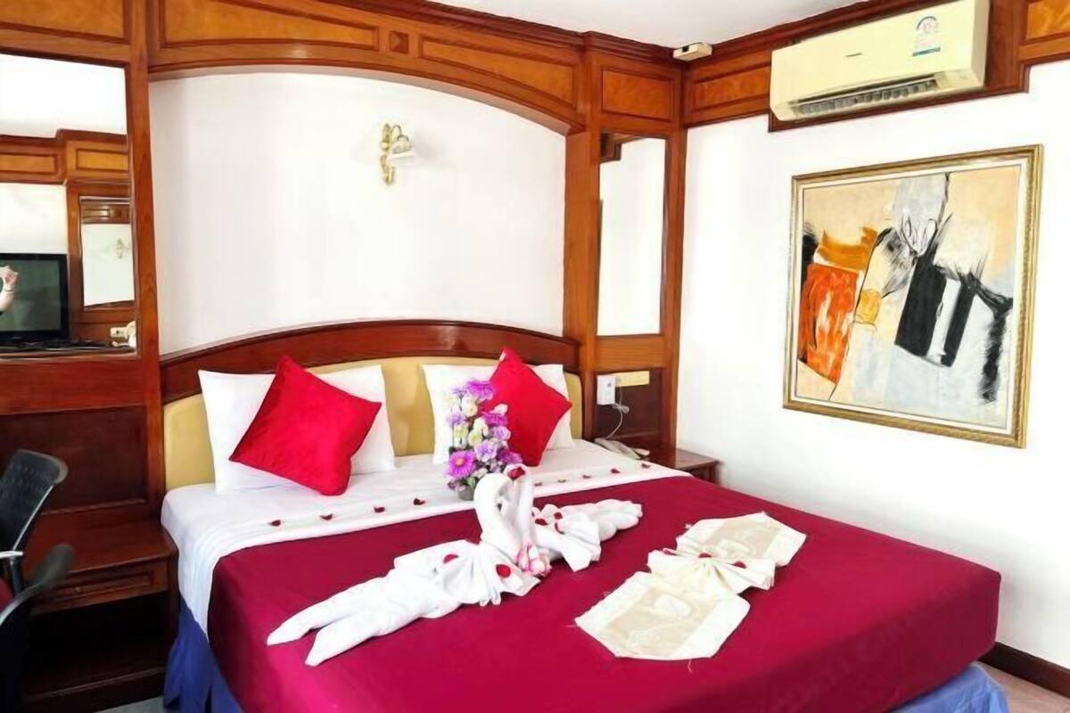 A hotel room with a bed, a wooden headboard, a painting on the wall, and a television in the corner.