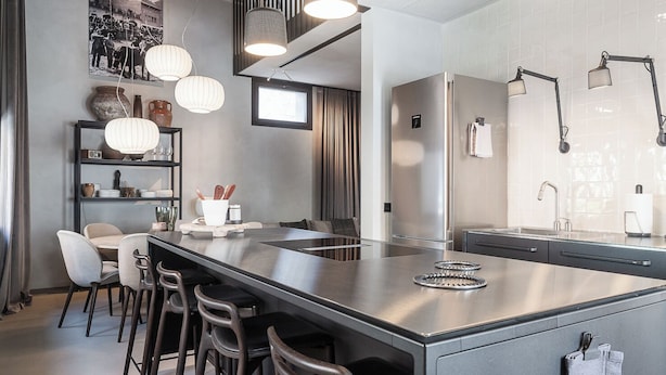 Una cocina moderna con una isla central, taburetes de bar, iluminación colgante y un refrigerador de acero inoxidable.