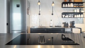 Una cocina moderna con un refrigerador de acero inoxidable, un horno empotrado y una encimera negra. Hay dos lámparas colgantes sobre el lavabo y una cafetera en la encimera.