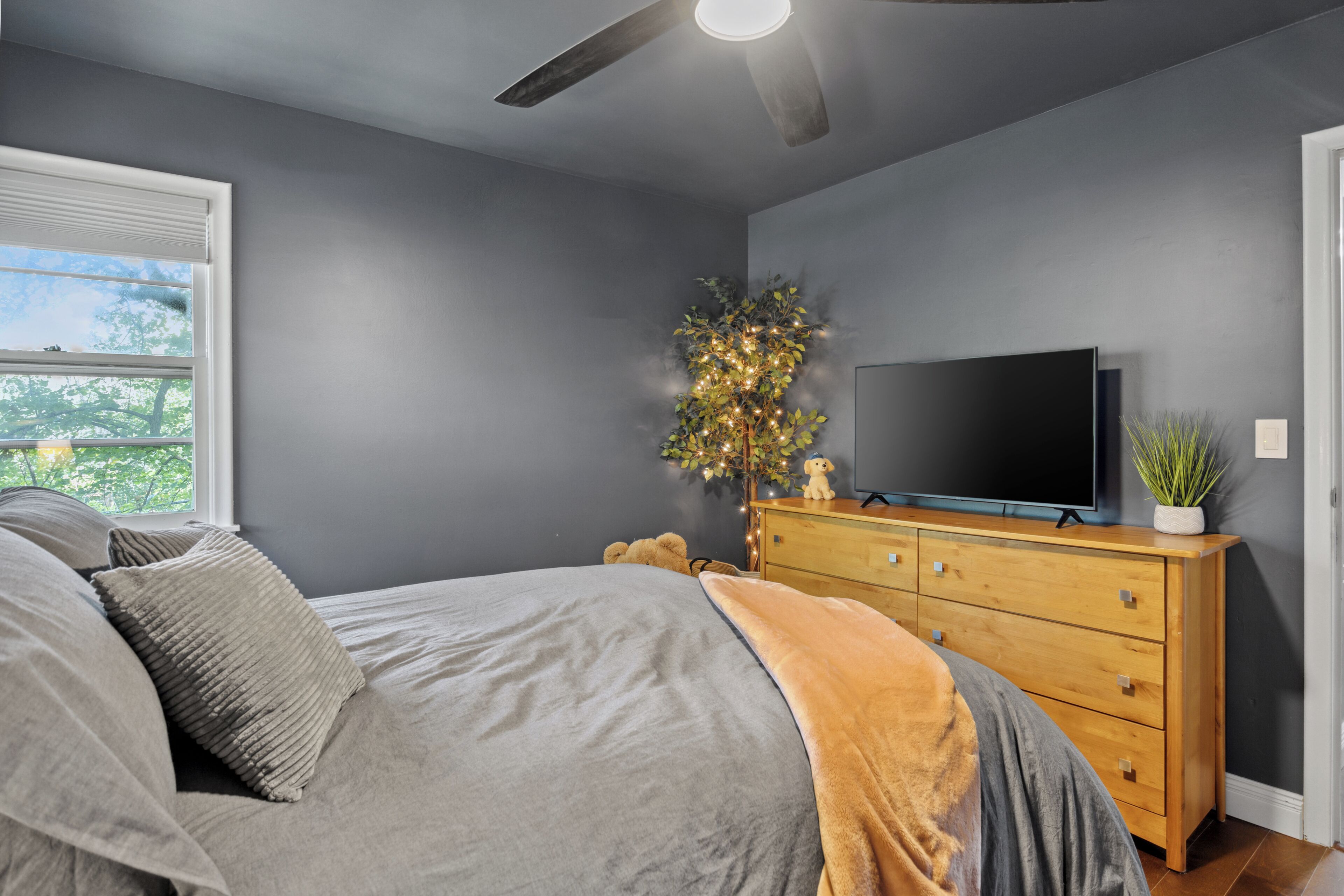 Une chambre à coucher comprenant un lit, une commode en bois, un téléviseur et une fenêtre avec des stores.