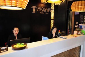 Una recepción de hotel con dos empleados, una pared negra con letras doradas y una lámpara colgante moderna.