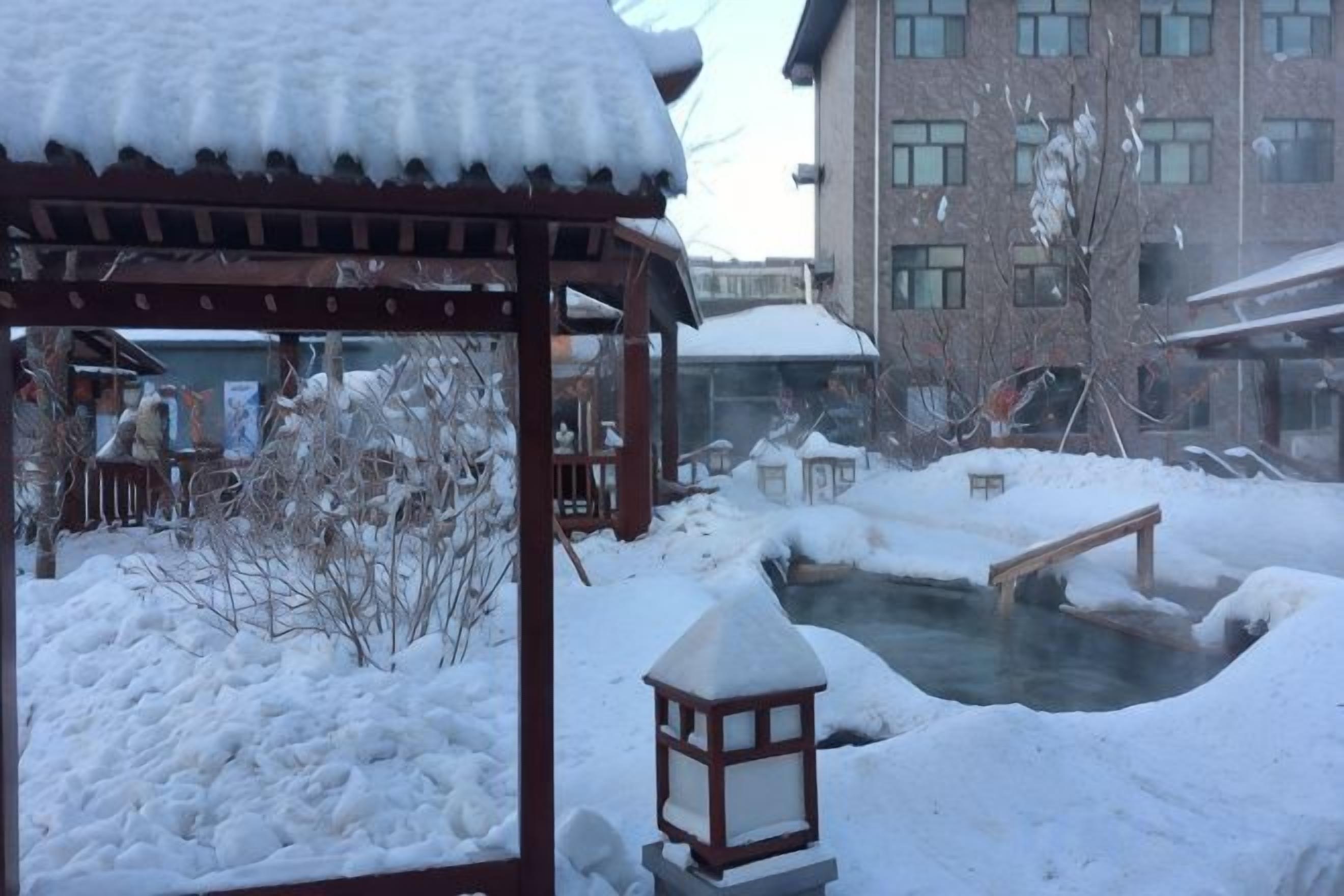 A snow-covered outdoor area with a wooden structure, a small pond, and a building in the background.