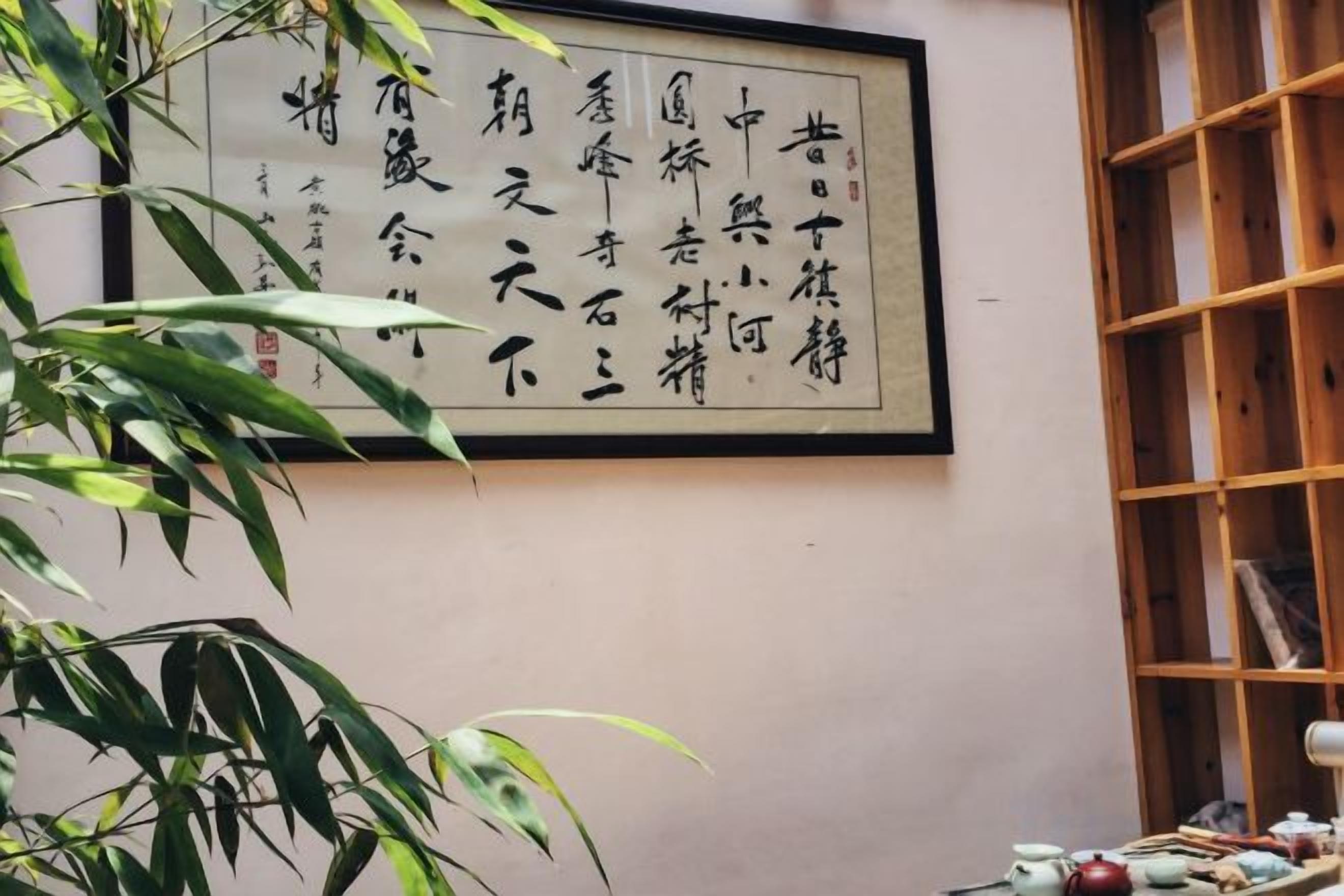 Ein Zimmer mit traditioneller chinesischer Kalligraphie an der Wand, einem hölzernen Bücherregal und einer Bambuspflanze im Vordergrund.