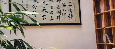 Ein Zimmer mit traditioneller chinesischer Kalligraphie an der Wand, einem hölzernen Bücherregal und einer Bambuspflanze im Vordergrund.