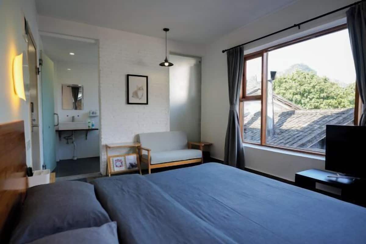 Un dormitorio con cama, una silla, televisión, ventana con cortinas, escritorio y espejo.