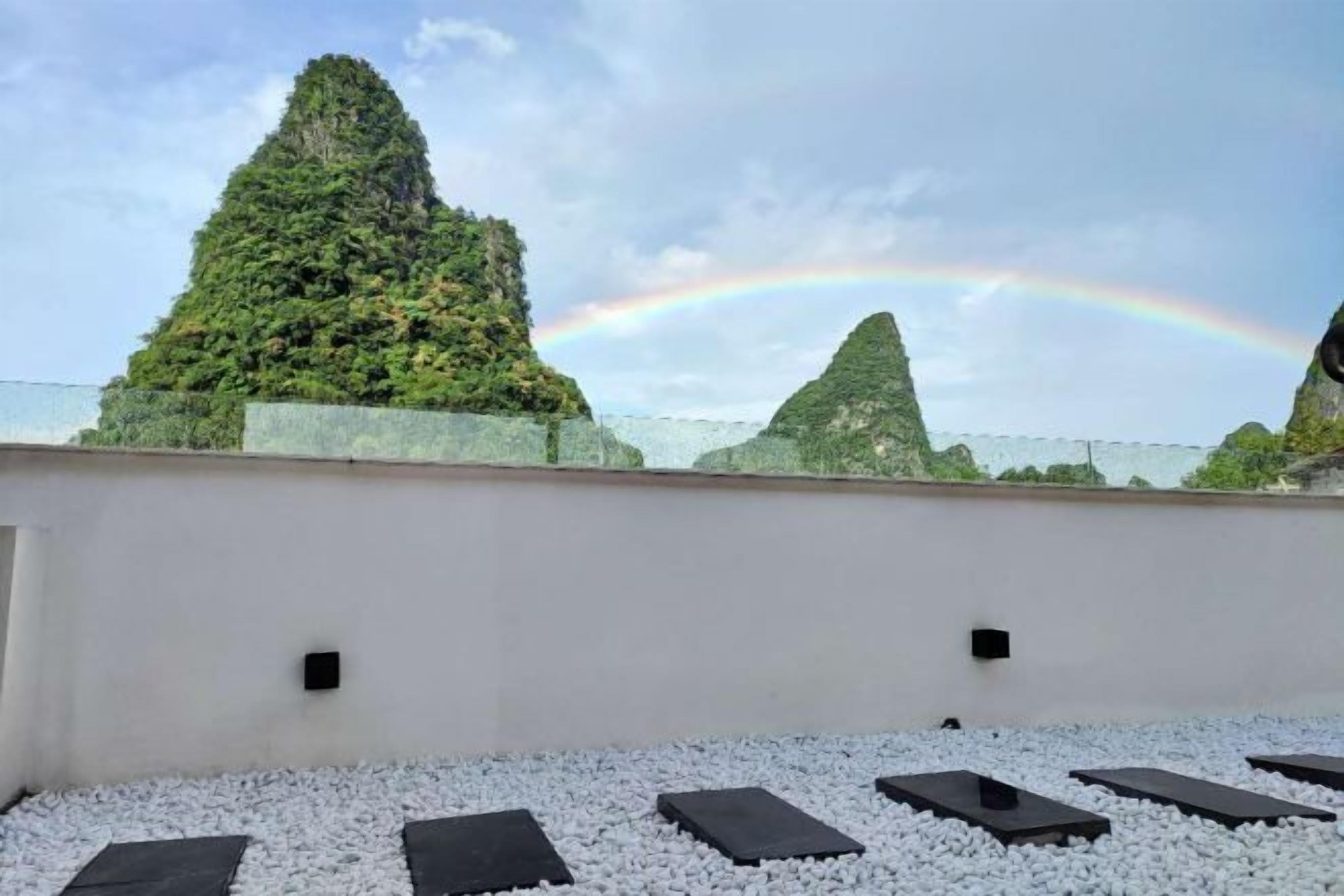 A rooftop terrace with a white wall, black rectangular planters, and a view of green hills with a rainbow.