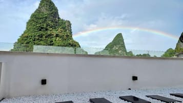 A rooftop terrace with a white wall, black rectangular planters, and a view of green hills with a rainbow.