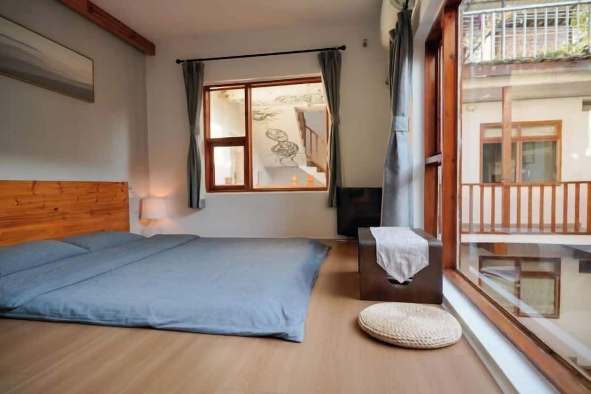 Un dormitorio con cabecera de madera, una cama, una mesita de noche con lámpara, una ventana con cortinas y vistas al balcón.
