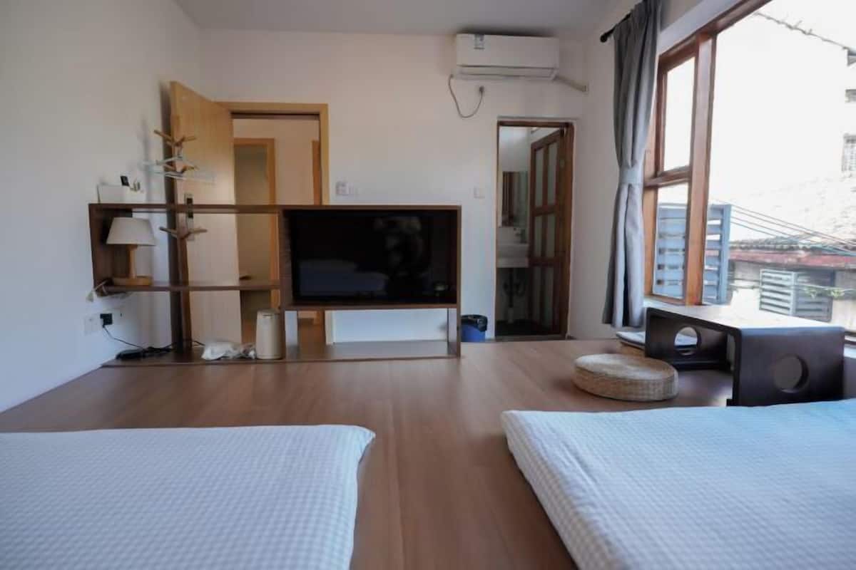Habitación de hotel con piso de madera, un televisor sobre un mueble bajo, una cama, un taburete redondo pequeño y un ventanal con cortinas.