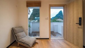 A room with a wooden floor, a single grey sofa, and a sliding door leading to an outdoor balcony with a view of a mountain and buildings. - Shifang Chuanshe Boutique Inn (Zhaoping)