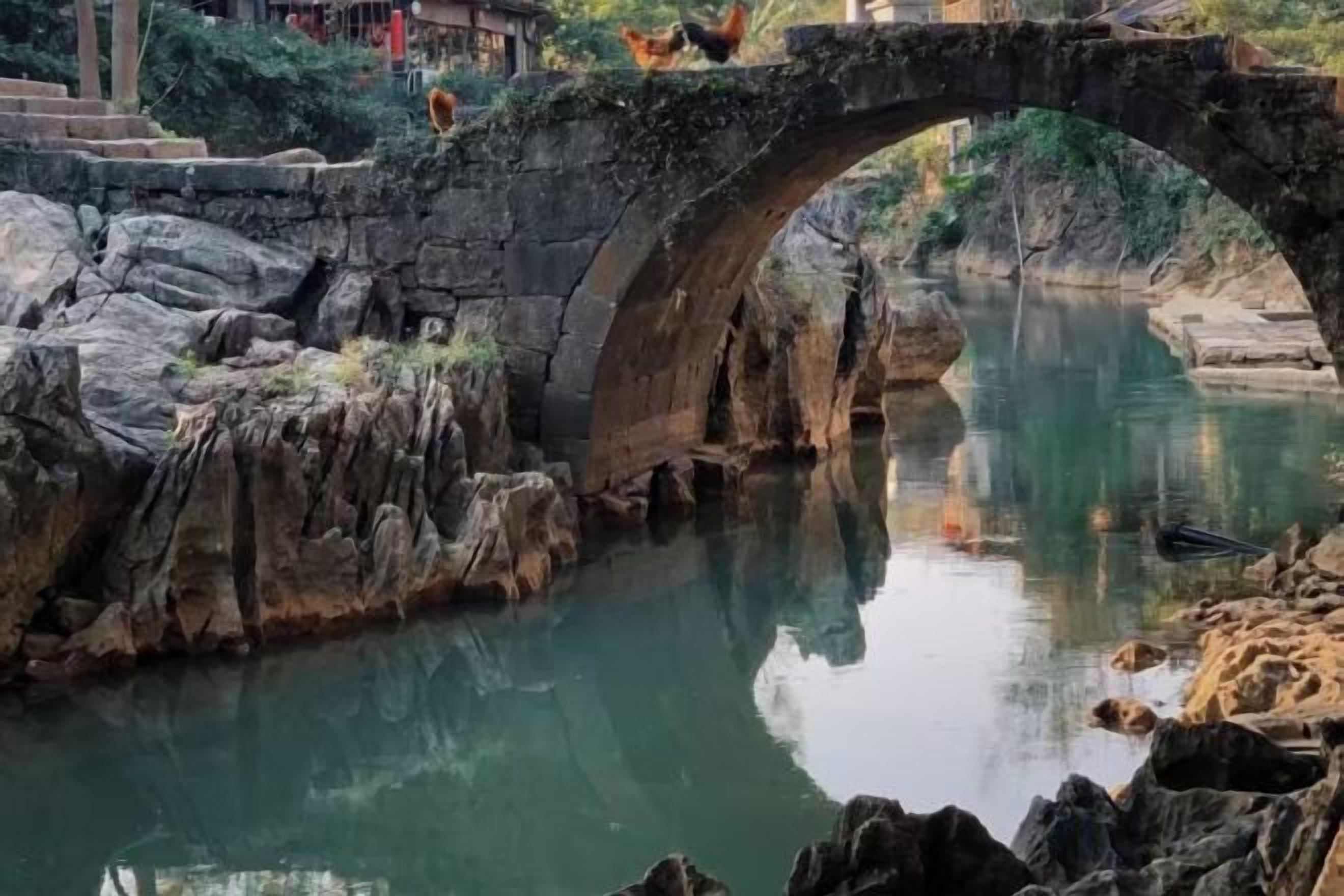 A stone bridge over a calm river with reflections, surrounded by rocks and vegetation.