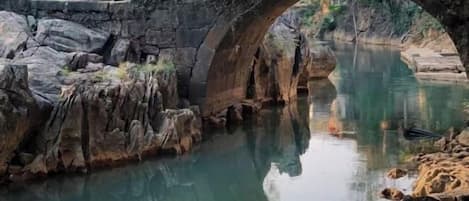 A stone bridge over a calm river with reflections, surrounded by rocks and vegetation.