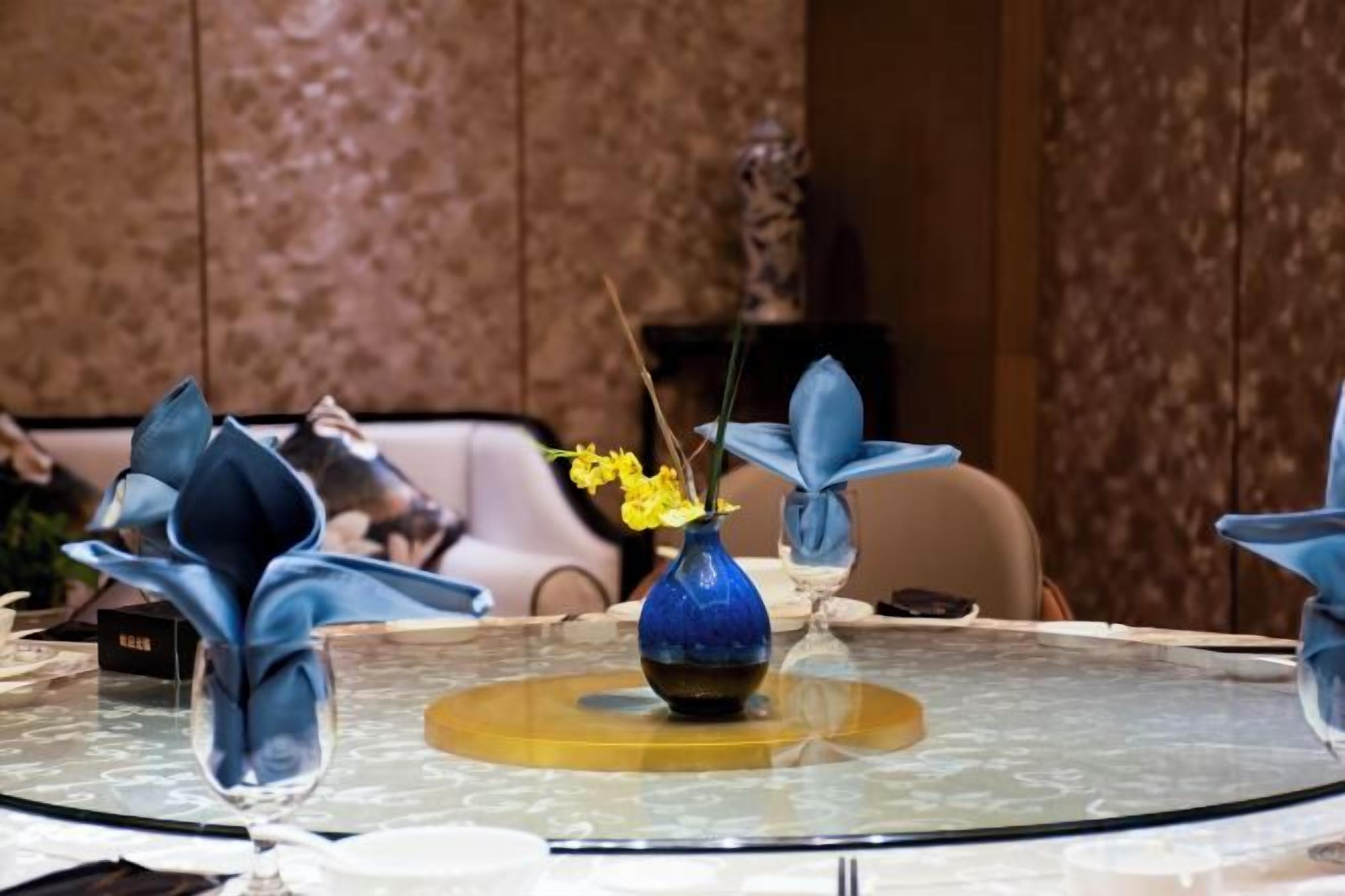 Una mesa redonda con un florero azul y flores amarillas, rodeada de servilletas dobladas.