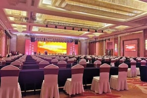 Une salle de conférence avec des rangées de tables et de chaises, une scène dotée d’un grand écran et un lustre.