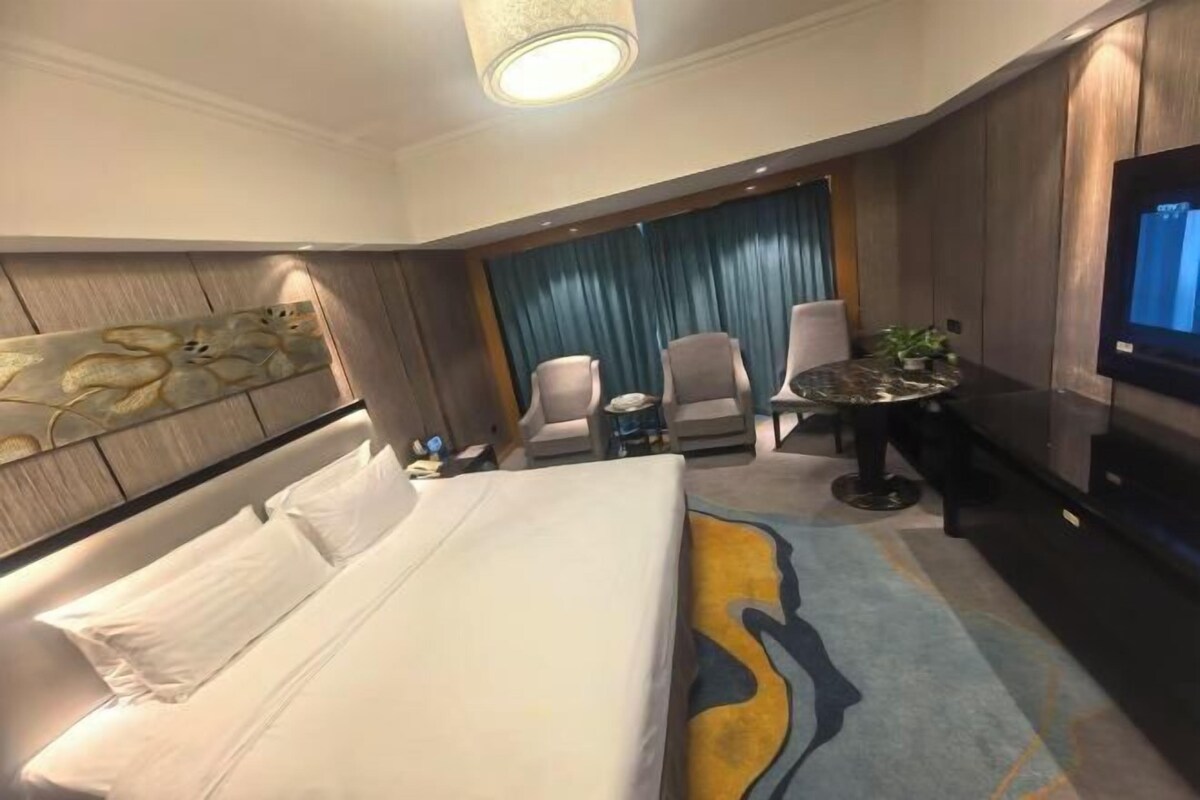 A hotel room with a bed, two armchairs, a small table, a TV, and a large window with curtains.