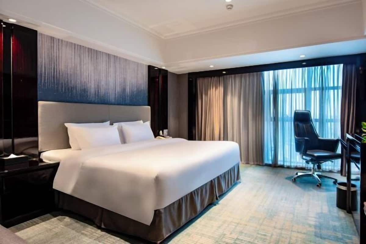 A hotel room with a large bed, a desk, a chair, and a view of the city through the window.
