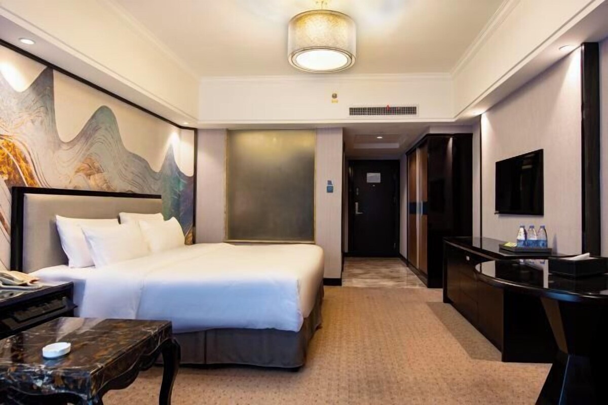 A hotel room with a bed, a desk, a chair, a television, and a painting on the wall.