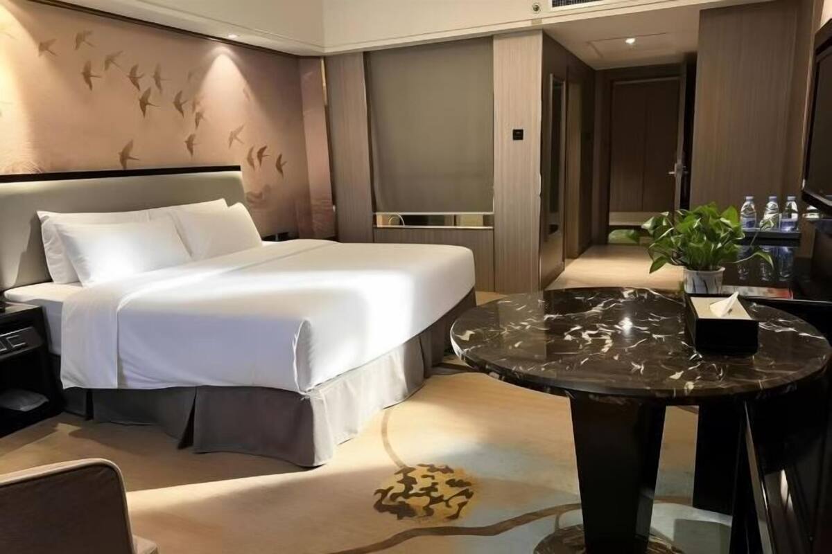 A hotel room with a bed, a round marble table, a nightstand, a wall-mounted TV, and a potted plant.