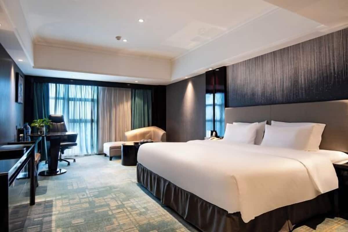 A hotel room with a large bed, a desk with a chair, a small sofa, and a large window with curtains.