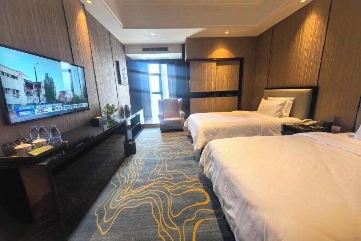 A hotel room with two beds, a TV, a desk, and a view of the outside.