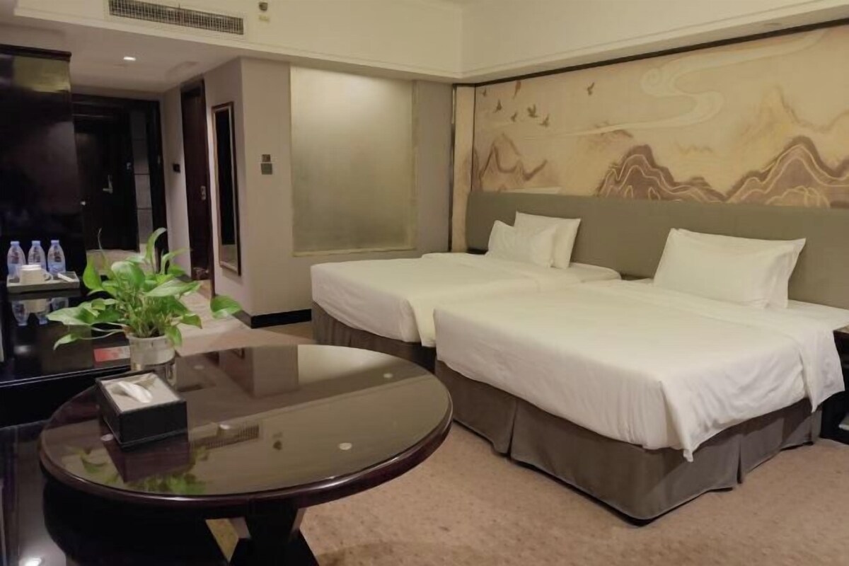 A hotel room with two beds, a round table, a potted plant, and a wall with a mountain design.