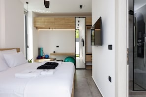 Quarto de hotel moderno com uma cama grande, TV fixada na parede, área de chuveiro e uma pequena cozinha.