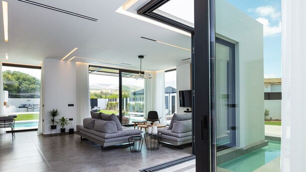 Ein modernes Wohnzimmer mit einem großen Glasfenster zum Pool, grauen Sofas und einem Essbereich mit Tisch und Stühlen.
