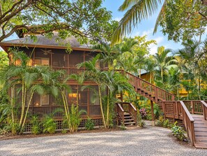 A two-story building with a wooden staircase leading to a balcony, surrounded by lush tropical vegetation.