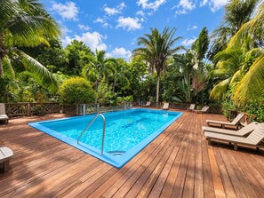 A wooden deck with a swimming pool, surrounded by lush greenery and palm trees.