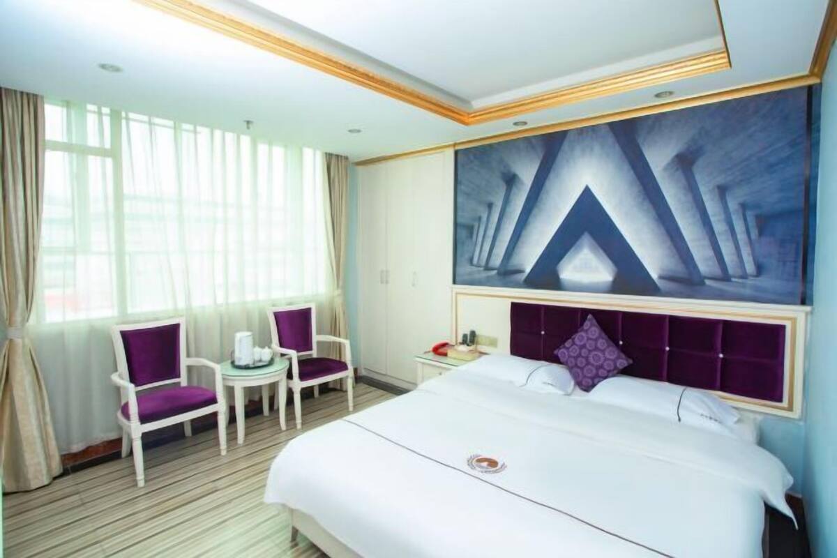 A hotel room with a large bed, a small table, two chairs, a nightstand, and a large abstract painting on the wall.