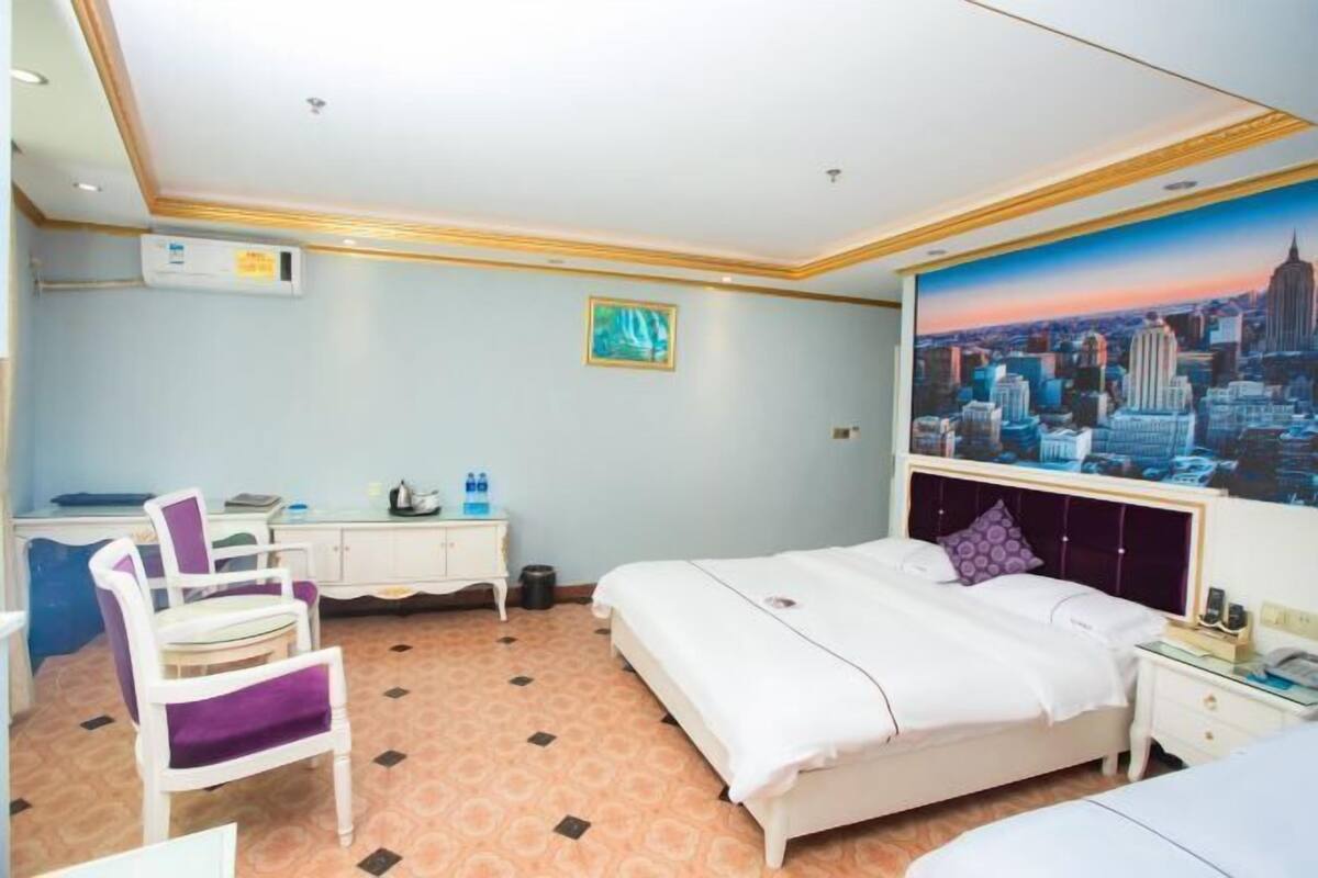 A hotel room with a bed, a desk, a chair, a window with a cityscape view, and an air conditioning unit.